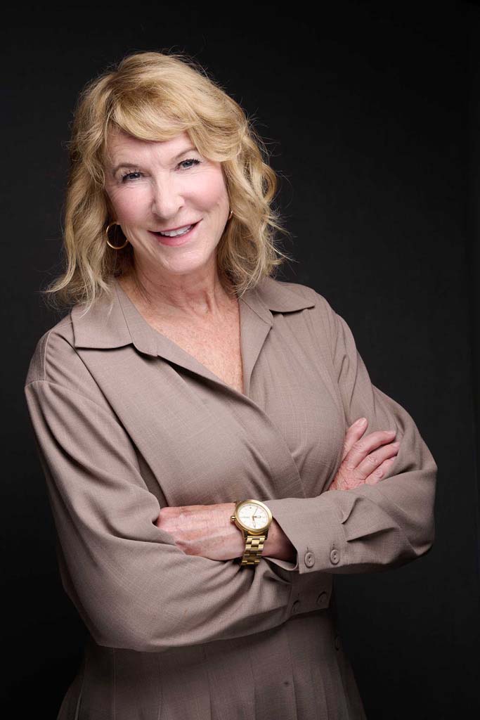 A blonde woman with her arms crossed in front of a black background.