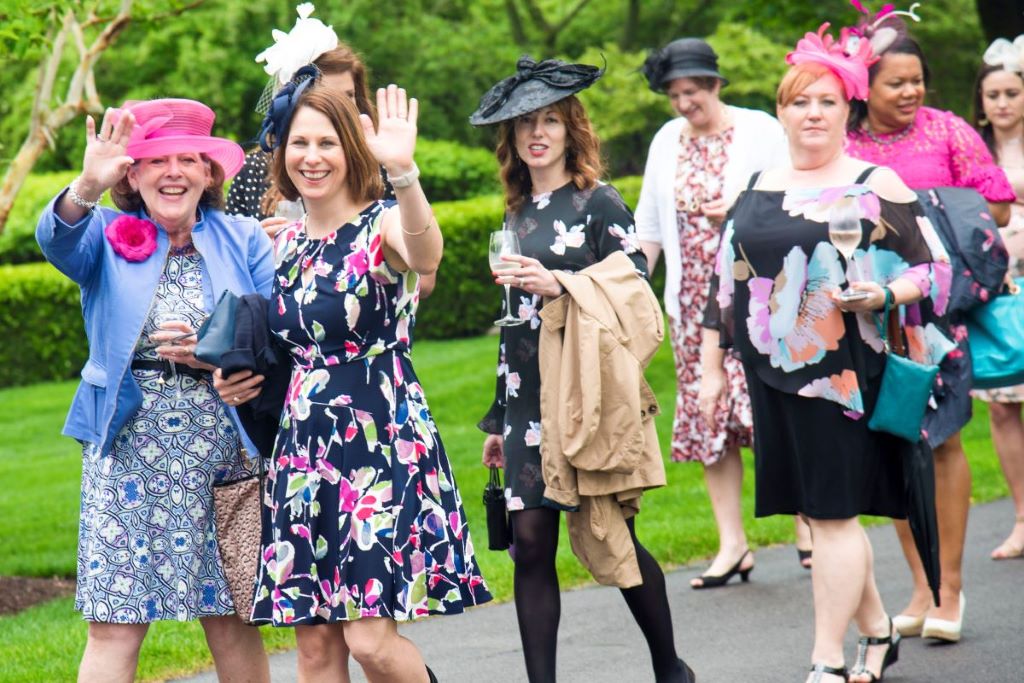 A group of women in dresses and hats wave at the camera